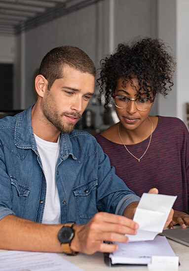 Young multiethnic couple checking bills while managing accounts on home banking app. Serious guy and african woman sitting at home discussing finance for the month. Young casual man and girl using laptop while looking at invoice and plan the budget to save.